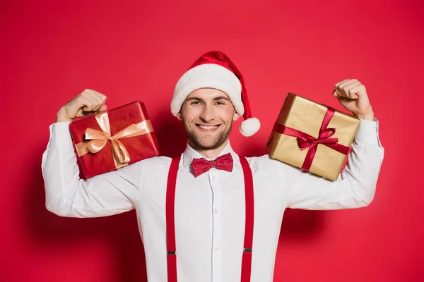 Hombre alegre en santa hat sosteniendo cajas de regalo sobre fondo rojo - foto de stock