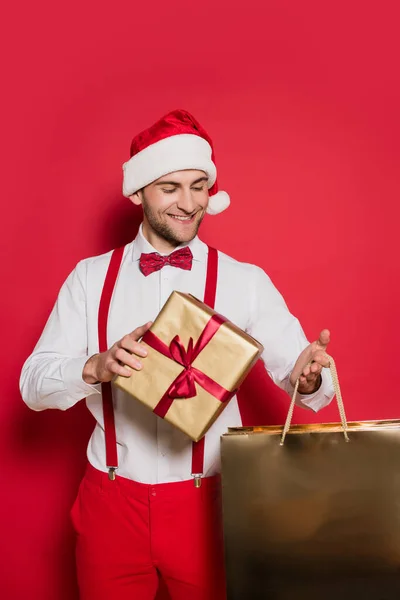 Cheerful man in santa hat holding shopping bag and present on red background — Stock Photo