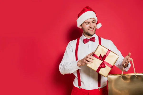 Homme positif dans le chapeau santa tenant boîte cadeau et sac à provisions sur fond rouge — Photo de stock