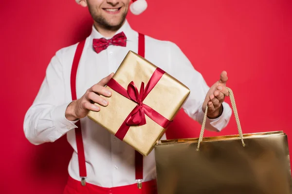 Cropped view of gift box and shopping bag in hands of smiling man blurred on red background — Stock Photo