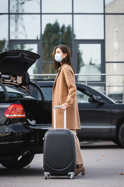 Elegante donna in abito autunnale e maschera medica guardando lontano mentre in piedi vicino al bagagliaio auto aperta con valigia — Foto stock