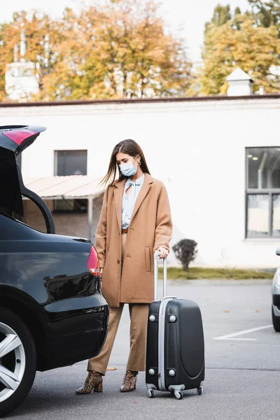 Donna in maschera medica ed elegante vestito autunnale in piedi vicino al bagagliaio auto aperta con valigia — Foto stock