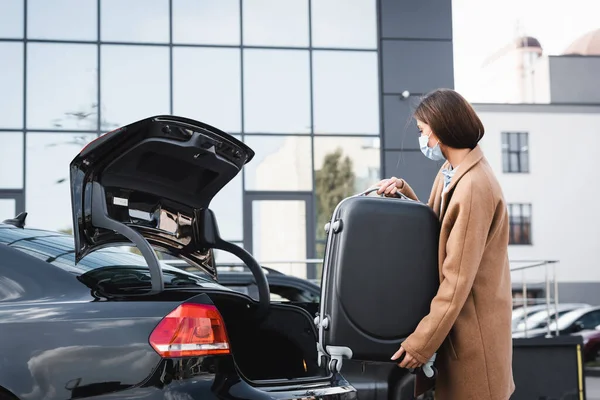 Young woman in medical mask loading suitcase in car trunk — Stock Photo