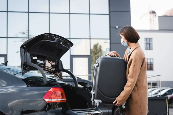 Jeune femme en masque médical chargement valise dans le coffre de la voiture — Photo de stock