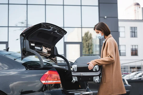 Mujer en gabardina y máscara médica poniendo maleta en el maletero del coche — Stock Photo