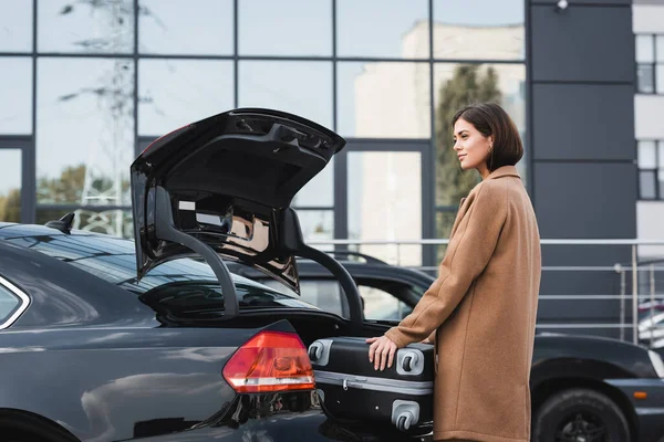 Donna sorridente in abito autunnale che distoglie lo sguardo mentre carica la valigia nel bagagliaio dell'auto — Foto stock