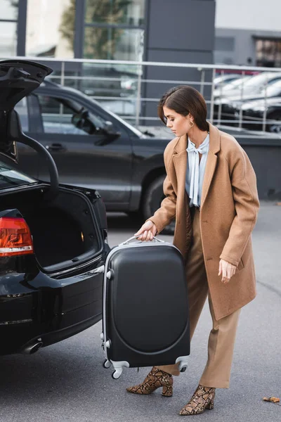 Стильна жінка в осінньому одязі тримає валізу біля багажника відкритого автомобіля — стокове фото