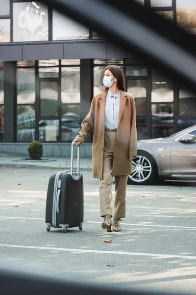 Jovem mulher em roupa de outono elegante e máscara médica andando ao longo do estacionamento do carro com mala em primeiro plano borrado — Fotografia de Stock