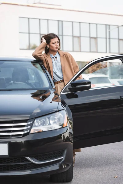 Verträumte Frau im schicken Trenchcoat lehnt an schwarzem Auto und schaut weg — Stockfoto