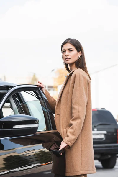 Молода жінка в стильному тренч пальто дивиться далеко під час відкриття дверей автомобіля — стокове фото