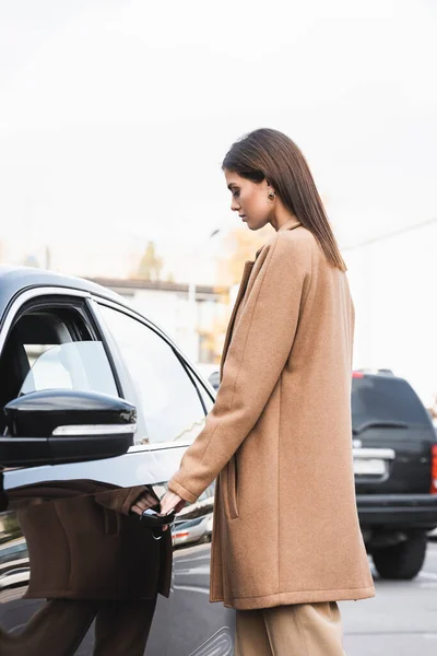 Junge Frau im schicken Trenchcoat öffnet Tür zu schwarzem Auto — Stockfoto