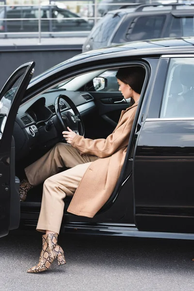 Mujer joven en traje elegante sentado en el coche con la puerta abierta y sosteniendo el volante - foto de stock