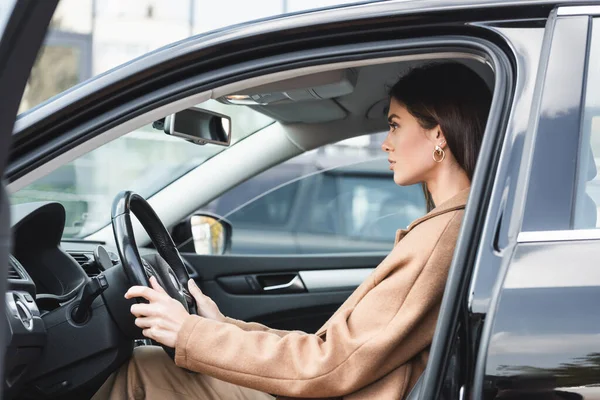 Stylische Frau im Trenchcoat sitzt im Auto mit offener Tür und hält Lenkrad — Stockfoto