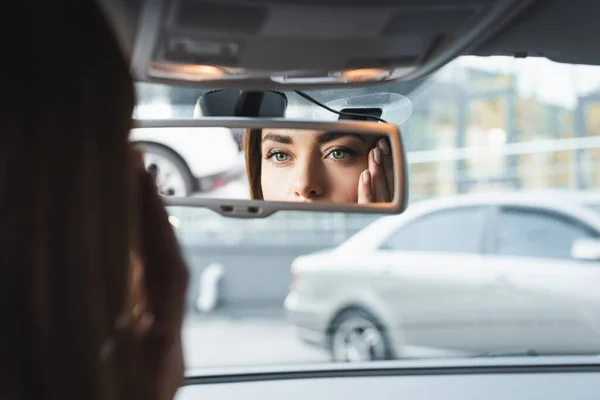 Mulher no carro tocando rosto enquanto olha no espelho retrovisor em primeiro plano borrado — Fotografia de Stock