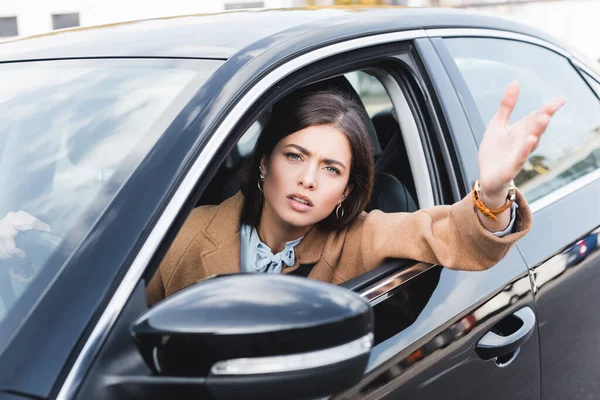 Mujer disgustada mirando por la ventana lateral y señalando con la mano mientras conduce el coche en primer plano borroso - foto de stock