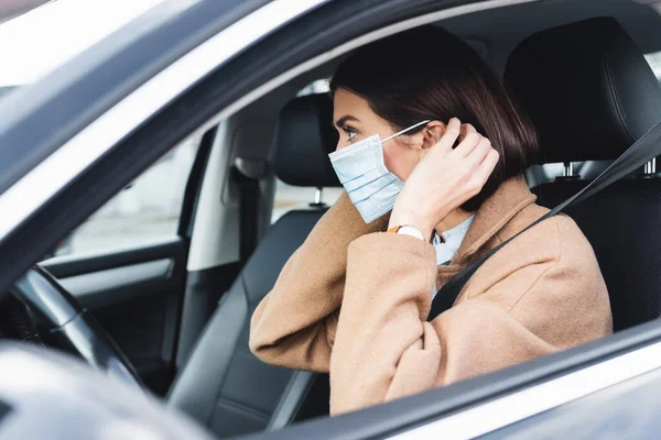 Jovem mulher no outono roupa colocando em máscara médica enquanto sentado no carro em primeiro plano borrado — Fotografia de Stock