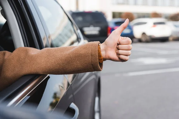 Vue recadrée de la femme montrant pouce vers le haut de la fenêtre latérale tout en conduisant la voiture sur le premier plan flou — Photo de stock