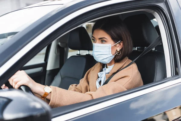 Mujer joven con máscara médica mirando hacia el futuro mientras conduce el coche en primer plano borroso - foto de stock