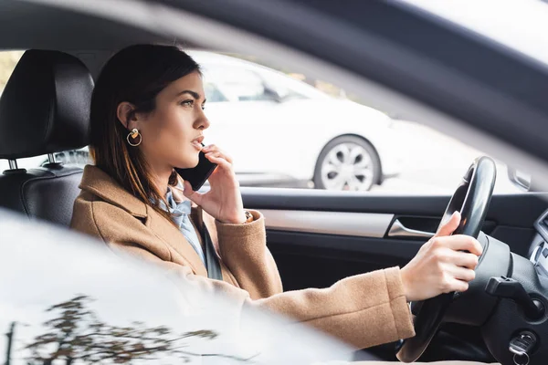 Vue latérale de la femme élégante en trench coat parlant sur téléphone portable tout en conduisant la voiture sur le premier plan flou — Photo de stock