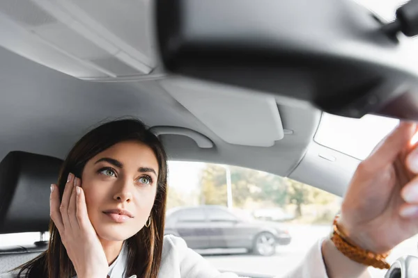 Jovem mulher tocando rosto enquanto olha no espelho retrovisor do carro em primeiro plano borrado — Fotografia de Stock