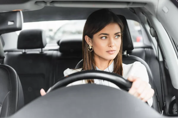 Jeune femme élégante regardant de côté tout en conduisant la voiture sur le premier plan flou — Photo de stock