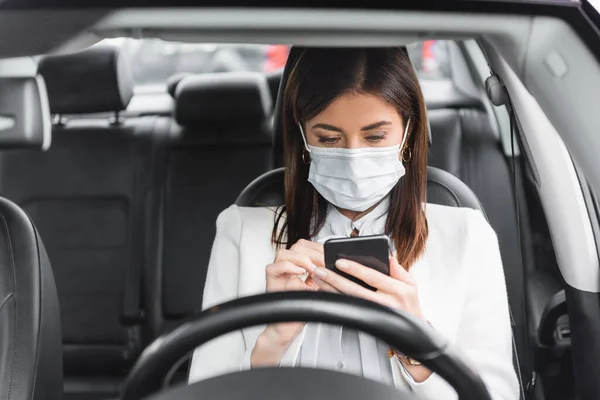 Young woman in medical mask messaging on smartphone in car on blurred foreground — Stock Photo