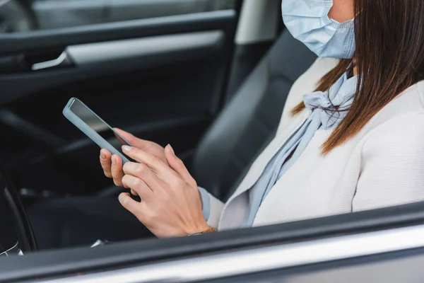 Vista recortada de la mujer en máscara médica escribiendo en el teléfono inteligente en el coche en primer plano borrosa - foto de stock
