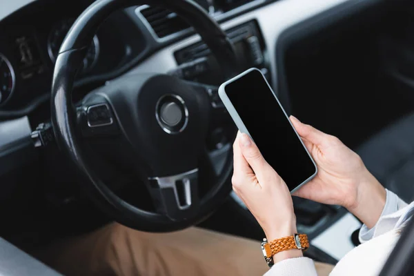 Vista parcial de la mujer en el reloj de pulsera que sostiene el teléfono inteligente con pantalla en blanco cerca del volante sobre fondo borroso - foto de stock