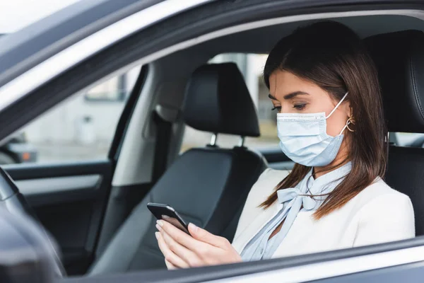 Femme élégante dans le masque médical bavarder sur le téléphone mobile en voiture sur le premier plan flou — Photo de stock