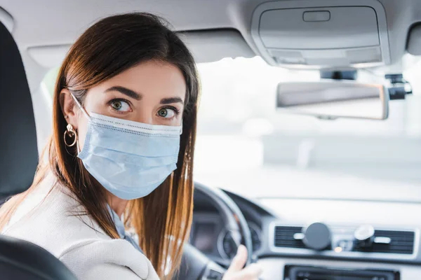 Mujer joven con máscara médica mirando hacia atrás mientras conduce el coche sobre fondo borroso - foto de stock