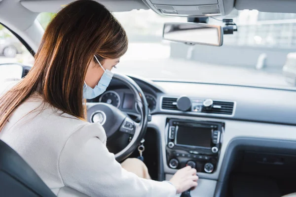 Jeune femme en masque médical levier de vitesse de déplacement tout en étant assis dans la voiture — Photo de stock