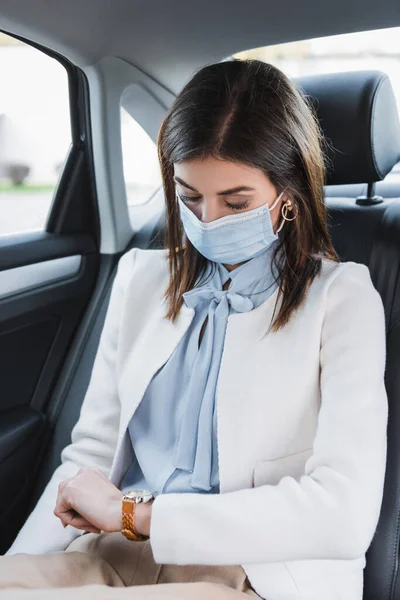 Stilvolle Frau mit medizinischer Maske schaut auf Armbanduhr, während sie im Auto auf dem Rücksitz sitzt — Stockfoto