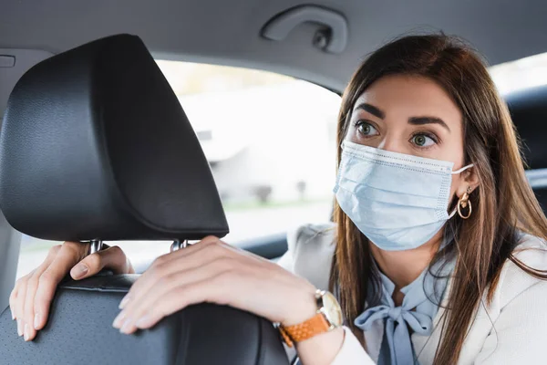 Young woman in medical mask touching front seat and looking ahead while riding in car — Stock Photo