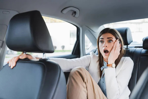 Scared woman holding hand on face while sitting on back seat of car on blurred foreground — Stock Photo