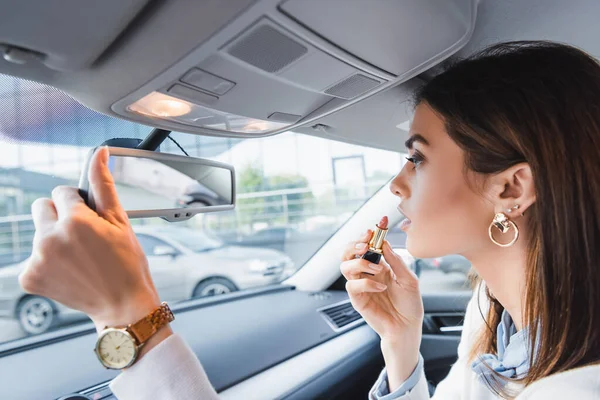 Mulher elegante segurando batom enquanto olha no espelho retrovisor do carro — Fotografia de Stock