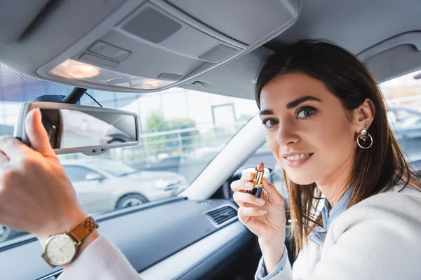 Mujer sonriente sosteniendo lápiz labial y mirando a la cámara mientras ajusta el espejo retrovisor en el coche en primer plano borroso - foto de stock