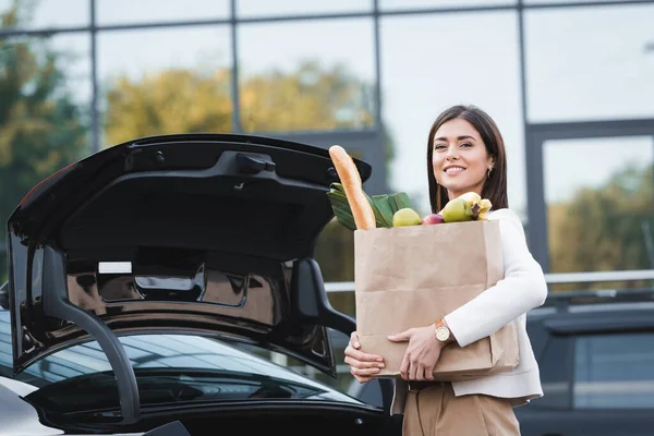 Femme heureuse regardant la caméra tout en se tenant près du coffre de voiture ouvert tout en tenant sac à provisions — Photo de stock