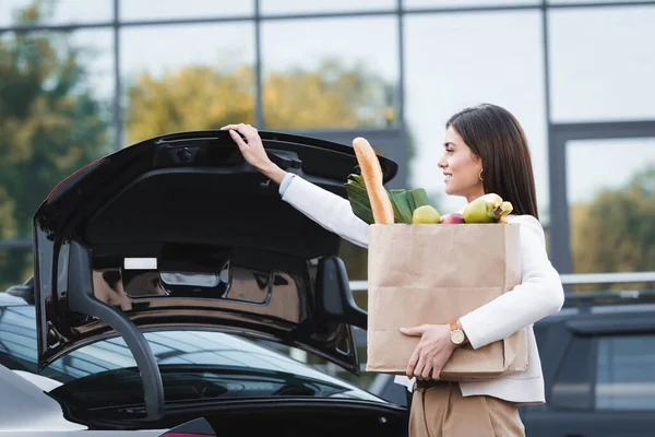 Femme souriante ouverture coffre de voiture tout en tenant sac à provisions avec de la nourriture — Photo de stock