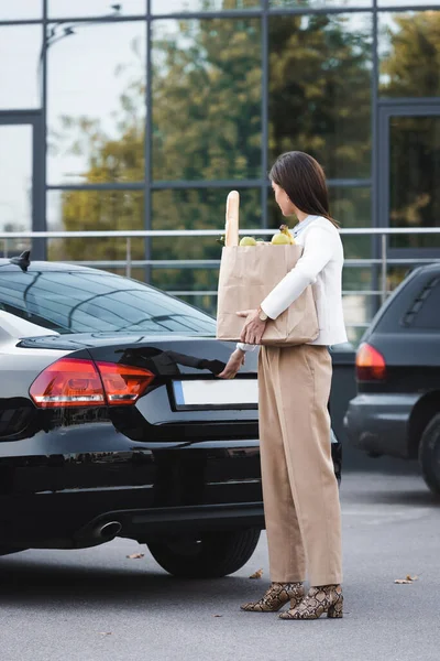 Mujer elegante apertura tronco de coche negro mientras sostiene bolsa de compras con comida - foto de stock