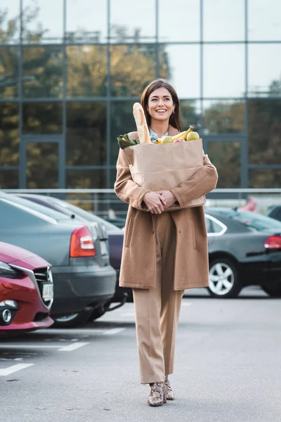 Mulher alegre em roupa de outono andando ao longo do estacionamento do carro enquanto carrega saco de compras com alimentos — Fotografia de Stock