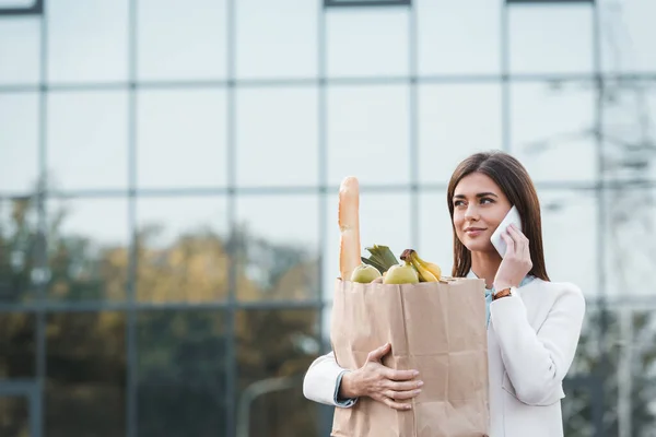 Mujer joven positiva mirando hacia otro lado mientras sostiene la bolsa de la compra con comida y hablando en el teléfono inteligente - foto de stock