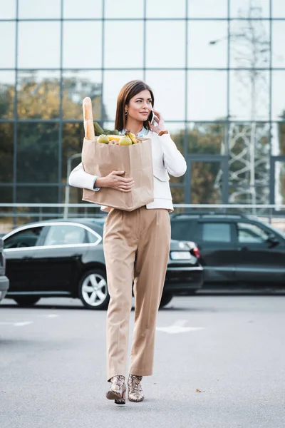 Mujer elegante hablando en el teléfono inteligente mientras camina a lo largo de aparcamiento con comida en la bolsa de compras - foto de stock