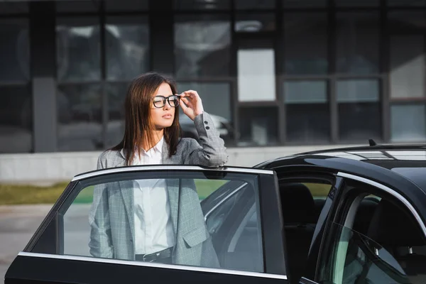 Stylish businesswoman touching eyeglasses while standing near open car and looking away — Stock Photo