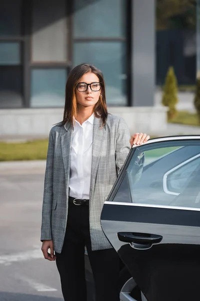 Young businesswoman in formal wear and eyeglasses standing near open car — Stock Photo