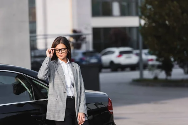 Sonriente mujer de negocios tocando gafas mientras está de pie cerca de coche negro - foto de stock