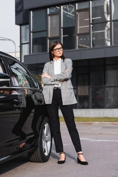 Giovane donna d'affari fiduciosa in piedi vicino auto nera con le braccia incrociate — Foto stock