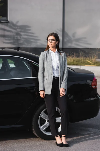 Молодая стильная деловая женщина в очках смотрит в сторону, стоя рядом с черной машиной — стоковое фото