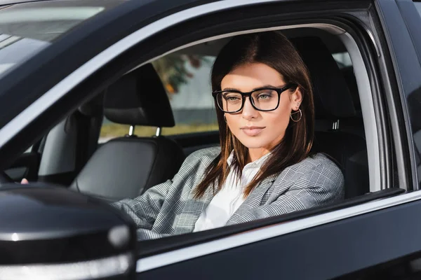 Stylish businesswoman in eyeglasses driving car on blurred foreground — Stock Photo