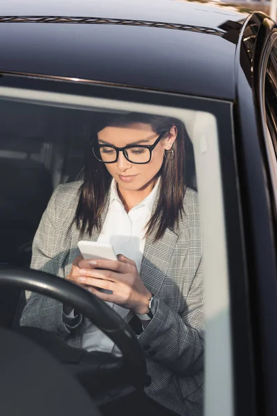 Stylish businesswoman in eyeglasses messaging on smartphone in car on blurred foreground — Stock Photo