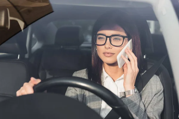 Стильна бізнес-леді в окулярах кермового автомобіля під час розмови на мобільному телефоні на розмитому передньому плані — стокове фото